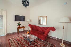 Una sala de estar con un sofá rojo, una mesa de centro de vidrio, una alfombra estampada, un televisor, una lámpara y un espejo.