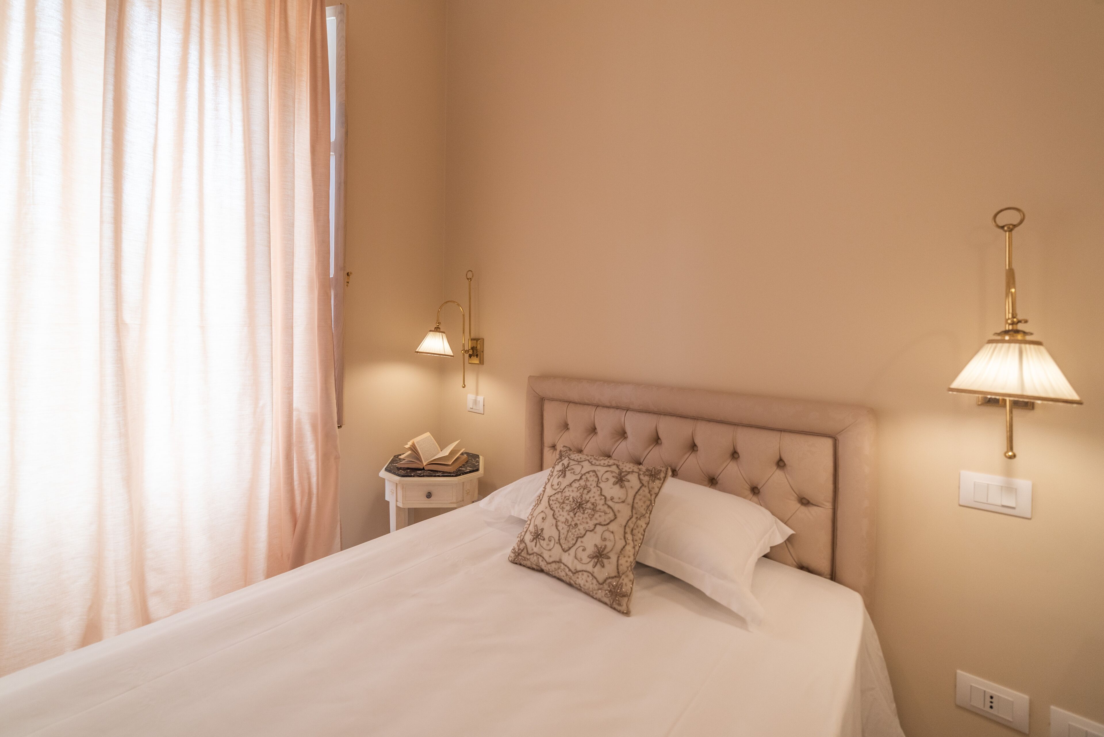 Een opgemaakt bed met een gestoffeerd hoofdeinde, een nachtkastje met een lamp en een raam met gordijnen.