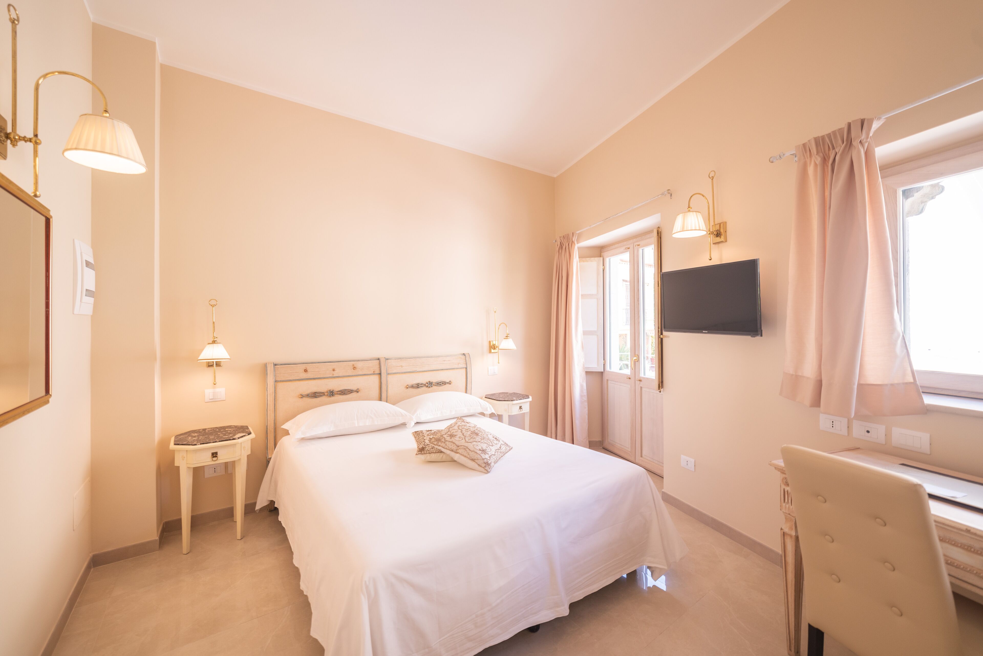A bedroom with a bed, bedside tables, a desk, a chair, a TV, and curtains.
