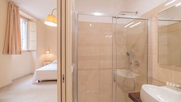 Un baño moderno con ducha envuelta en vidrio, una bañera grande y una mesita de noche con lámpara.