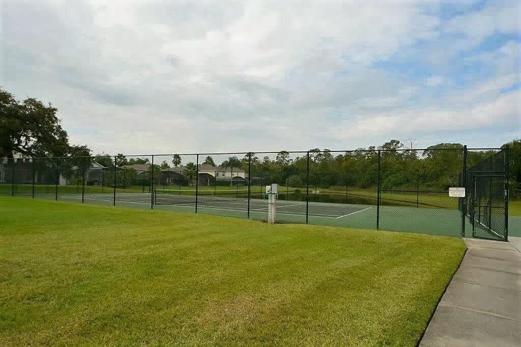 A tennis court with a fence, a grassy area, and a building in the background.
