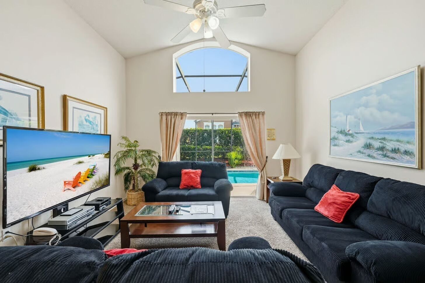 A living room with a TV, two sofas, a coffee table, a ceiling fan, and a view of a pool and greenery.