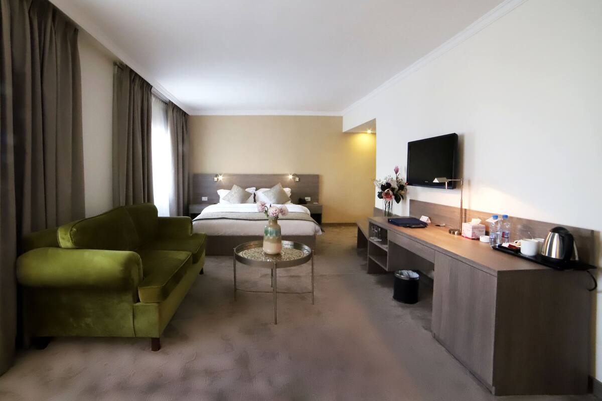 Ein Hotelzimmer mit Bett, einem grünen Sessel, einem kleinen runden Tisch mit einer Vase, einem Schreibtisch mit Flachbildfernseher und einer Kaffeemaschine.