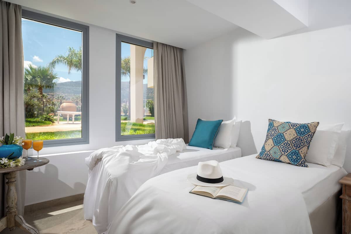 Una habitación de hotel con un ventanal grande, una cama con ropa de cama blanca, una mesita de noche con un libro y un sombrero, y vistas a una cúpula y vegetación.
