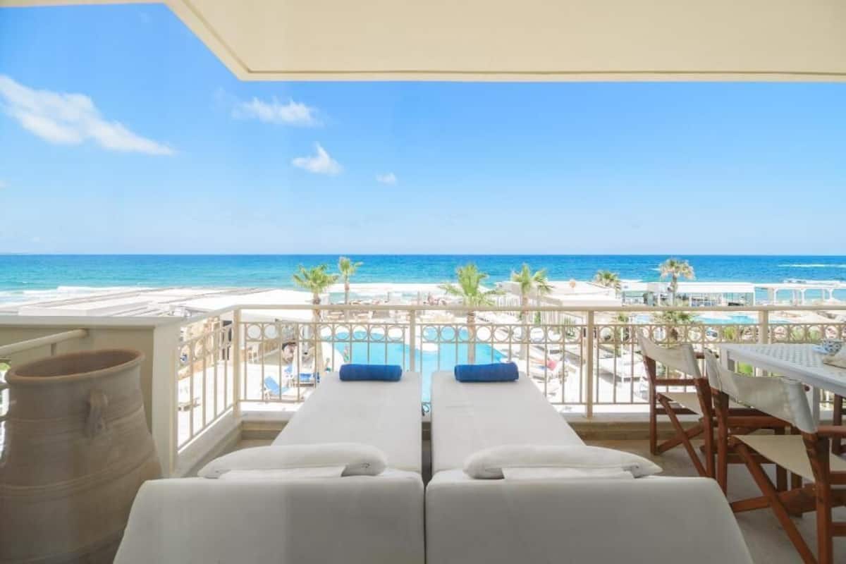 Un balcón con dos tumbonas blancas, una piscina y vistas a la playa y al océano.