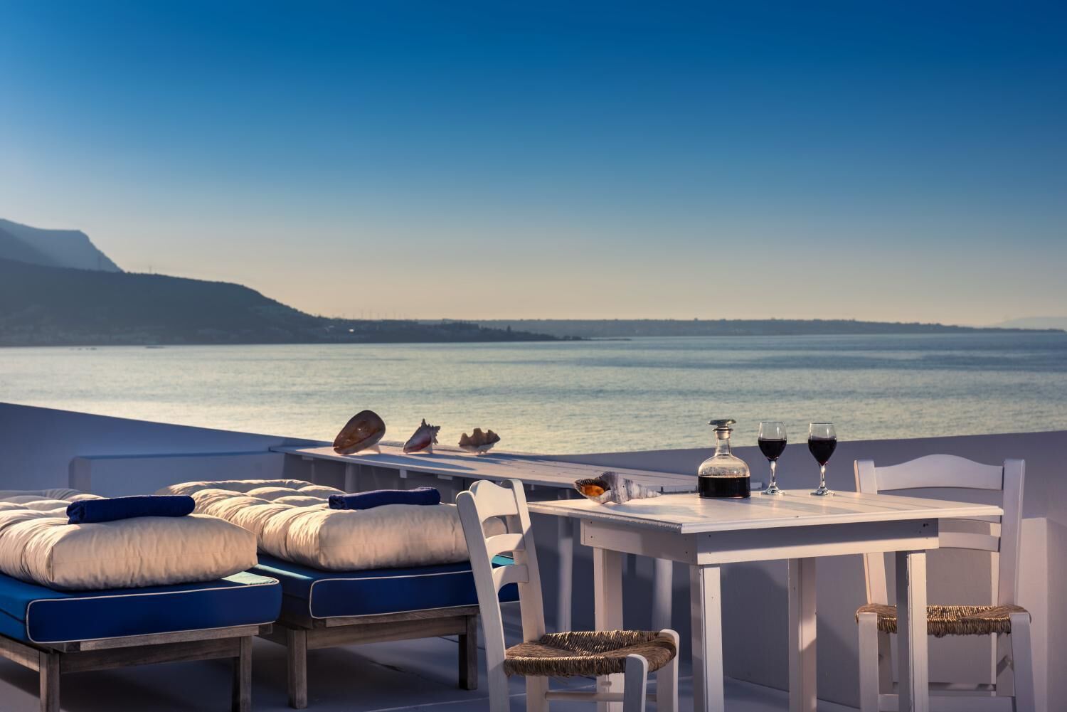 Una terraza con una mesa preparada para dos, con vista a un cuerpo de agua, y una cama y sillones de descanso al frente.