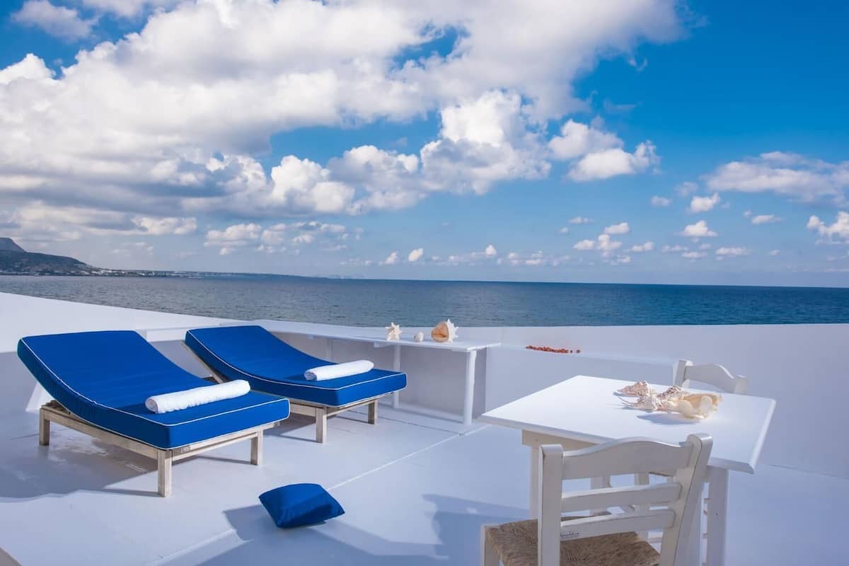 Una terraza iluminada por el sol con dos sillones azules, una mesa pequeña con mantel blanco y vistas al mar y a la costa lejana.