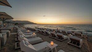 Un espace de détente aménagé au bord de la plage, avec des chaises longues et des tables, dont certaines sont décorées de bougies, offrant une vue sur l’océan.