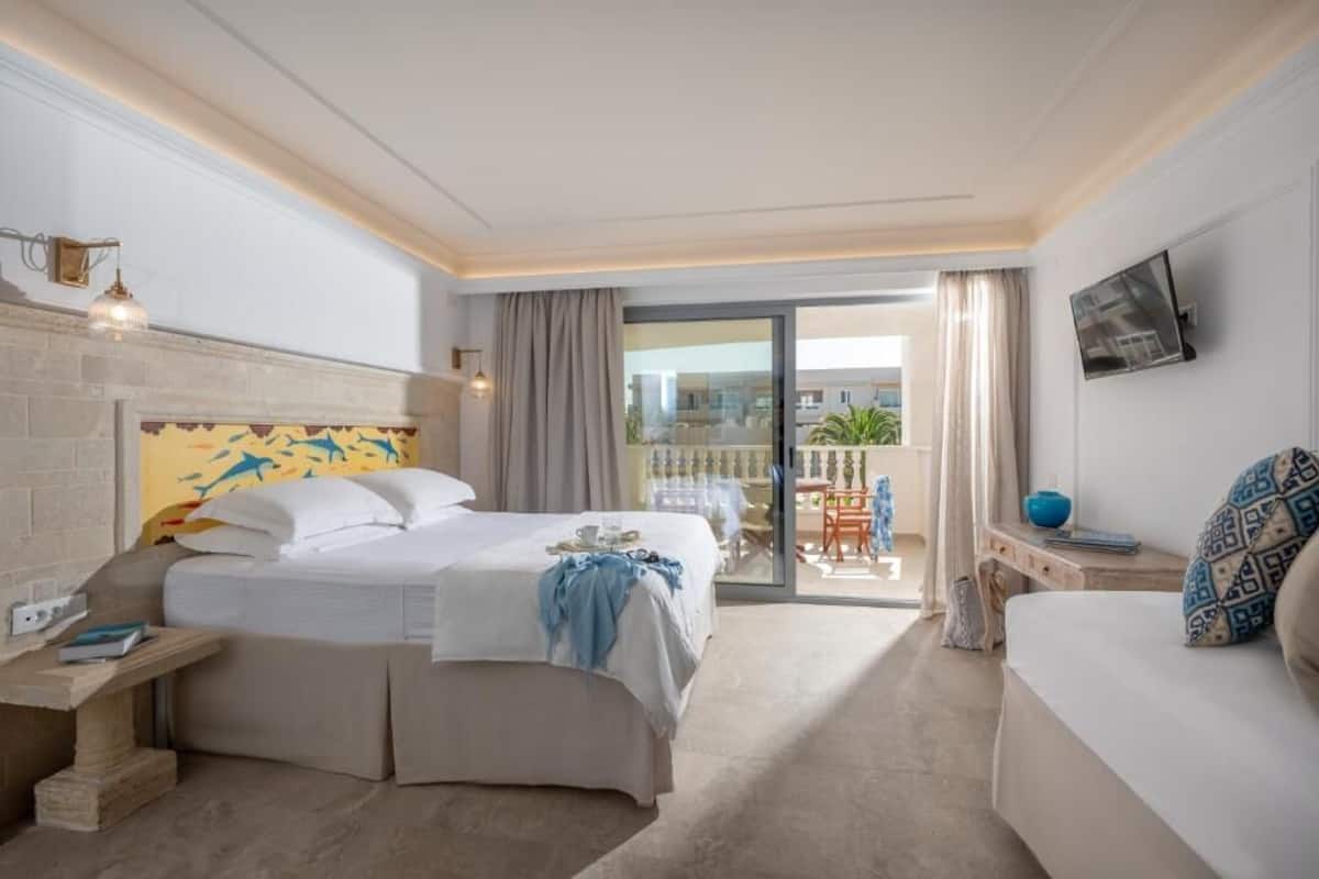 Habitación de hotel con cama, sofá, televisión, balcón con mesa y sillas, y vista a edificios.