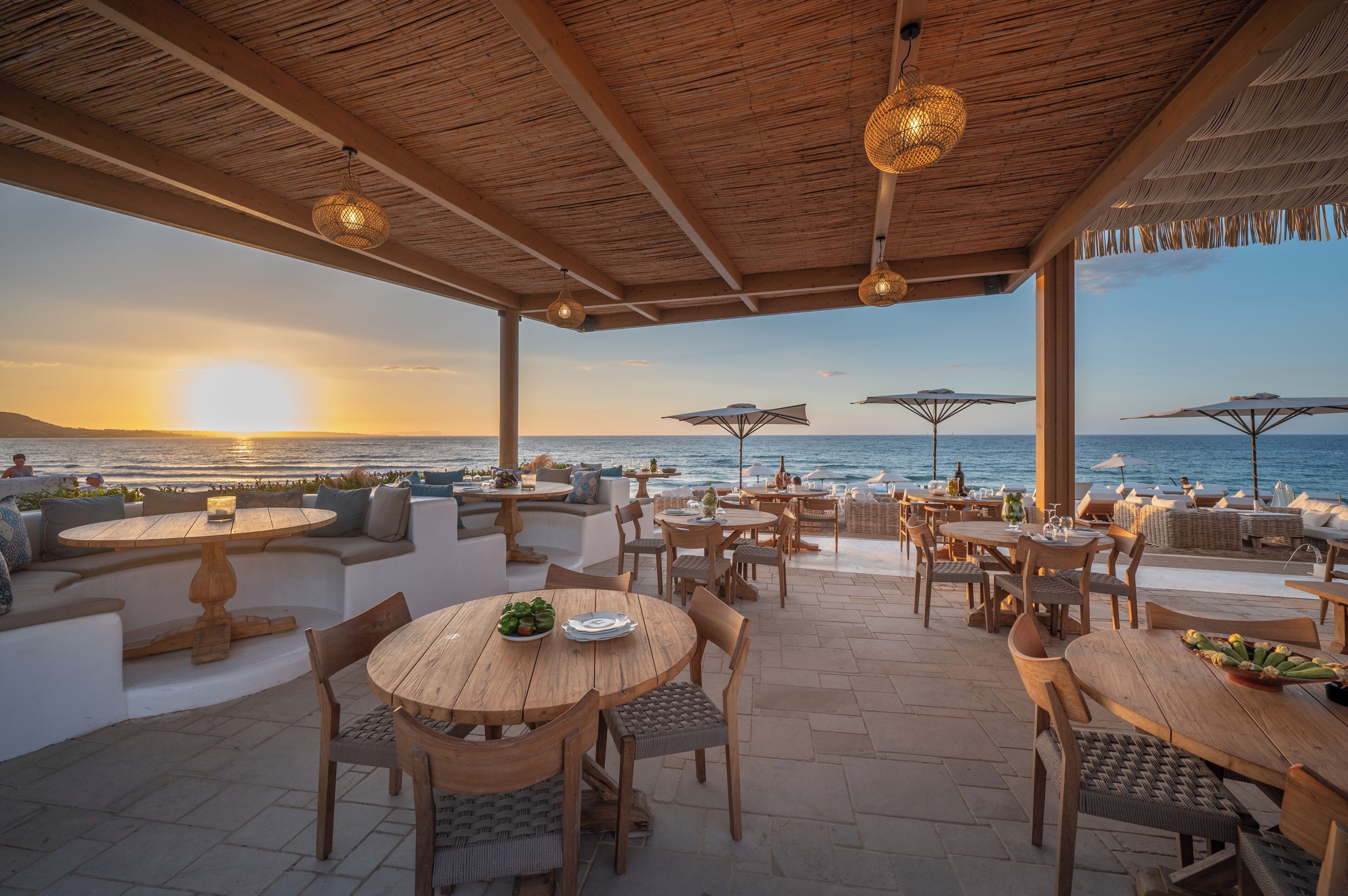 An outdoor seating area with wooden tables and chairs, umbrellas, and a view of the sea at sunset.