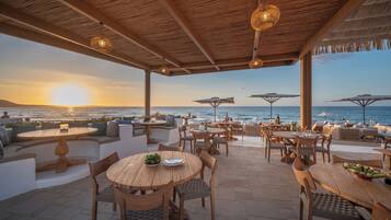 An outdoor seating area with wooden tables and chairs, umbrellas, and a view of the sea at sunset.