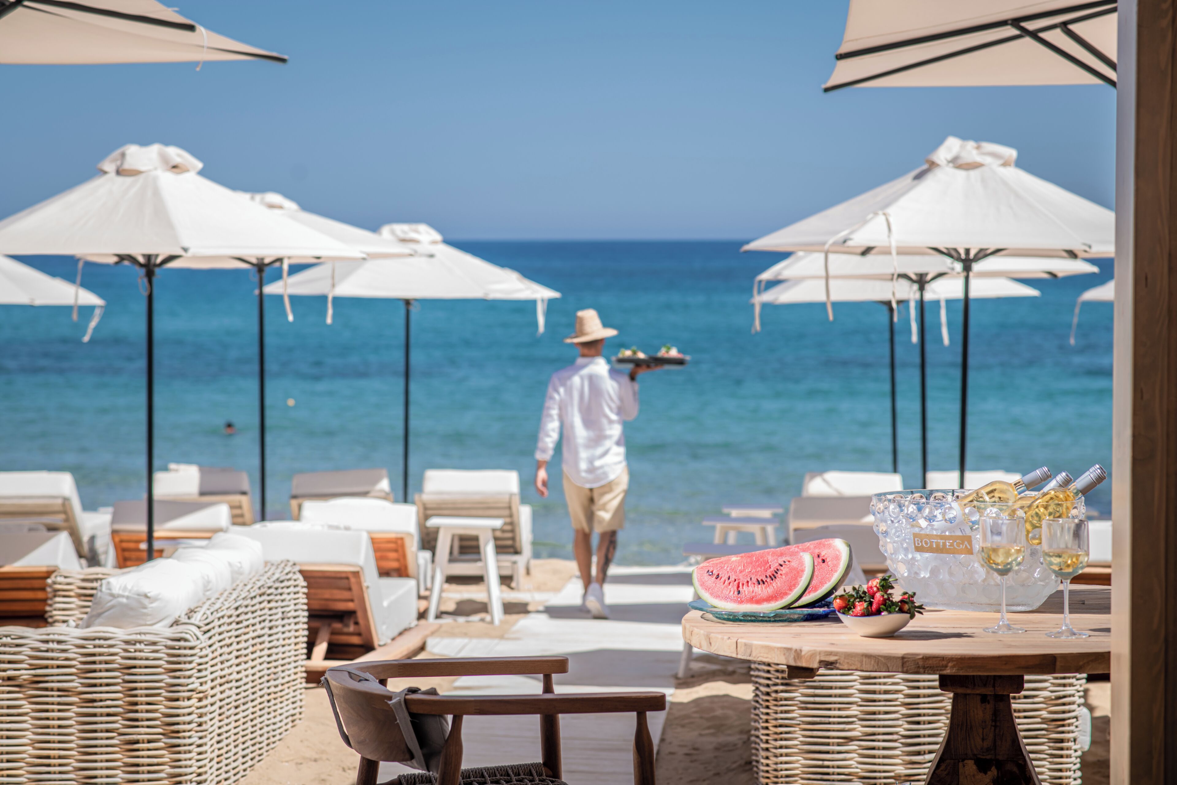 A beachside lounge with white umbrellas, wicker furniture, and a table with a watermelon and wine bottles.