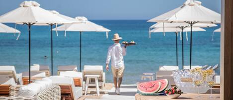 A beachside lounge with white umbrellas, wicker furniture, and a table with a watermelon and wine bottles.