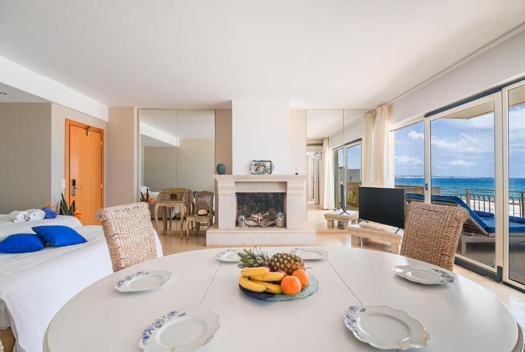 Habitación de hotel con cama, una mesa de comedor con fruta, chimenea y balcón con vista al mar.