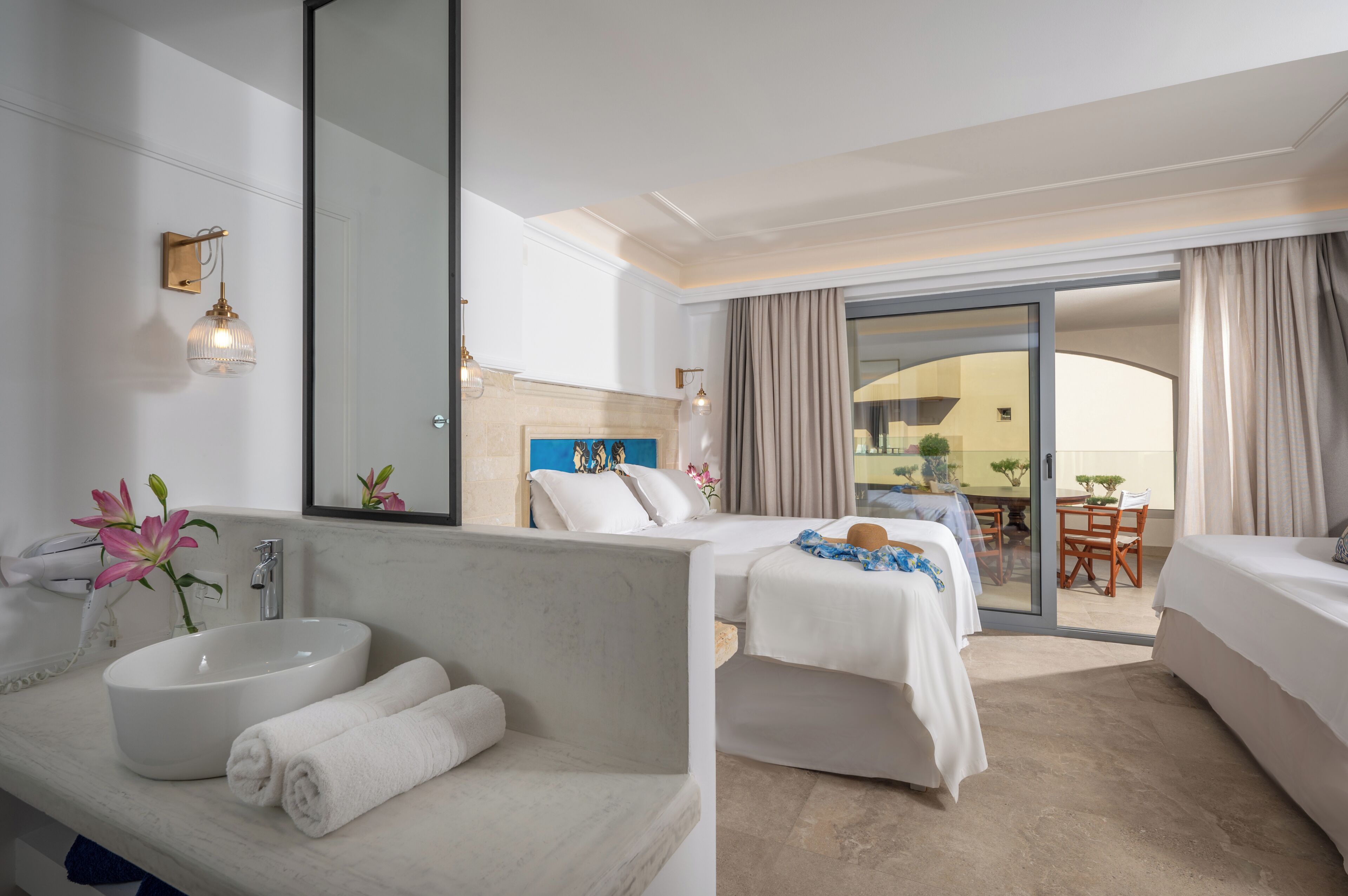 Habitación de hotel con una cama grande, un sofá independiente, un baño con lavamanos y espejo, y un balcón con mesa y sillas.