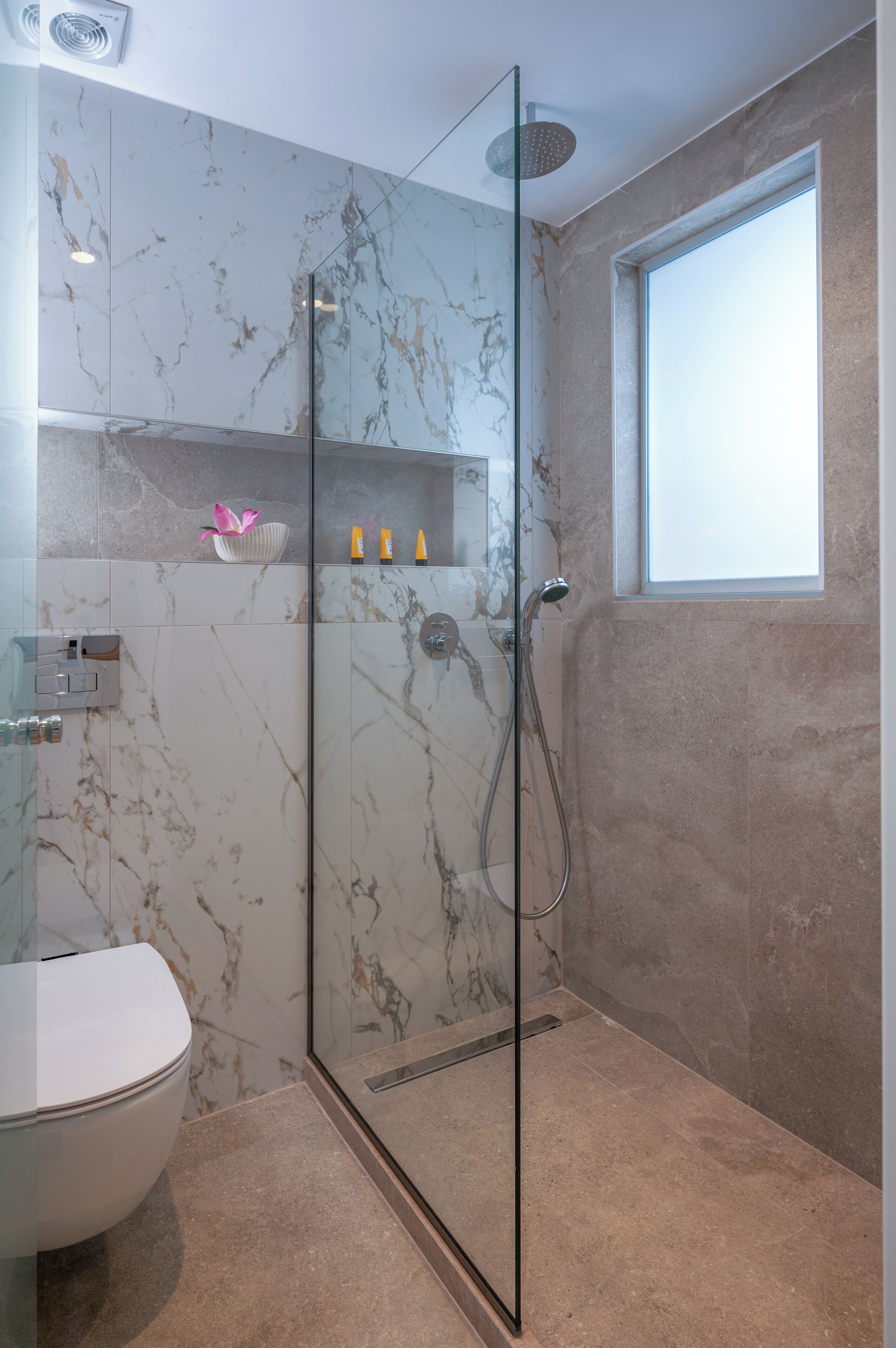 Een badkamer met een douchecabine van glas, een toilet, marmeren wanden en een raam.