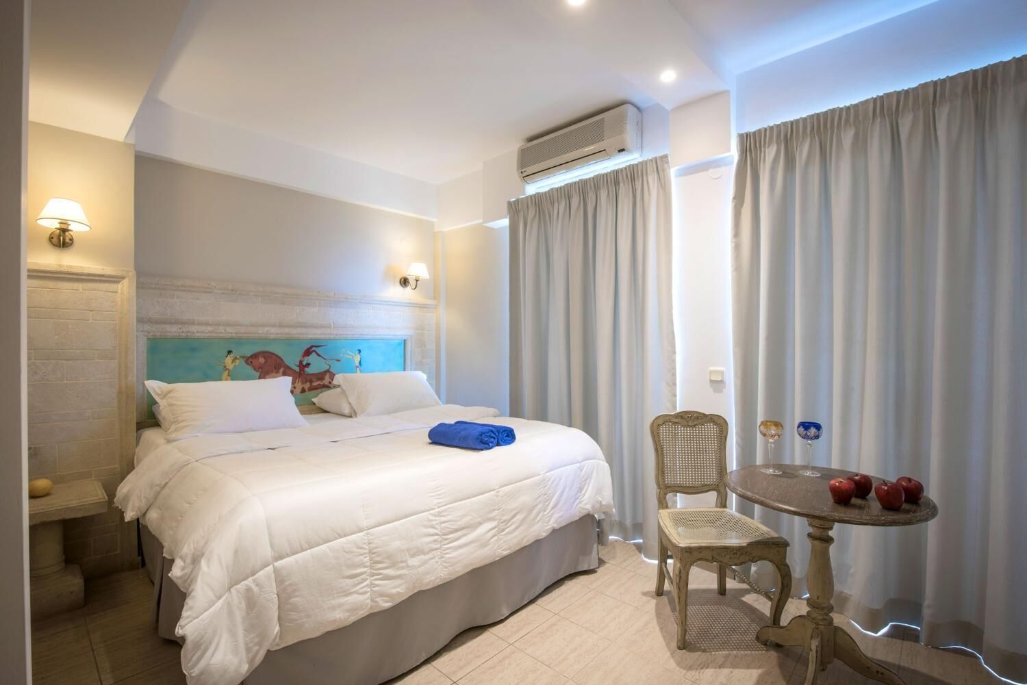 A hotel room with a bed, a small table with decorative items, a chair, and a wall-mounted air conditioner.