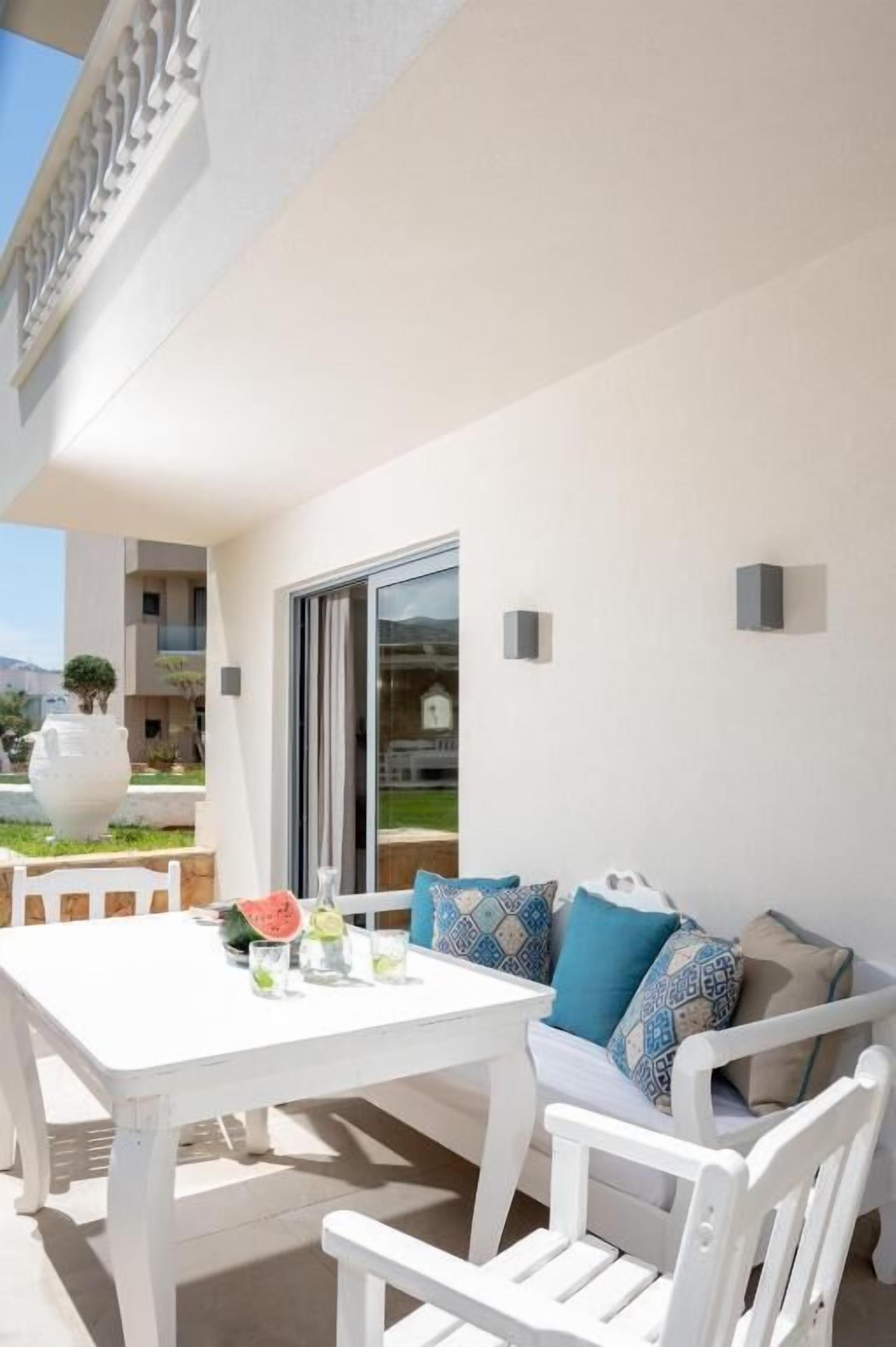 Un espace extérieur aménagé avec des sièges blancs, une table et des chaises, un mur blanc et une porte vitrée donnant sur un balcon.