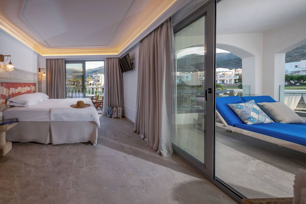 Habitación de hotel con cama, sofá, televisión y balcón con vistas a edificios y montañas.