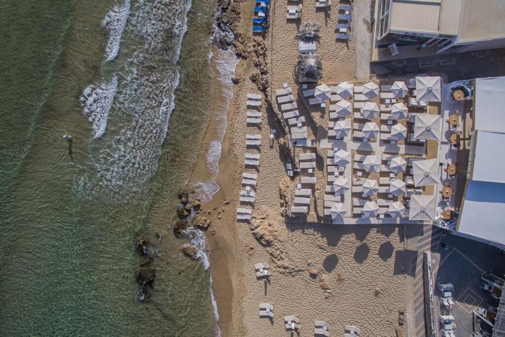 An aerial view of a beachside hotel with rows of sun loungers and umbrellas, a sandy beach, and a building adjacent to the shore.
