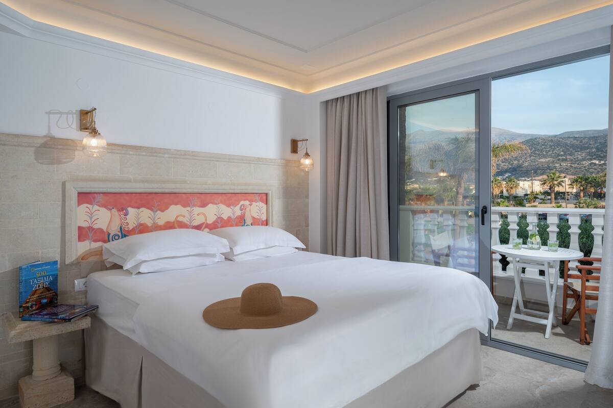 Un cuarto de hotel con una cama, una mesita de noche con un libro, un sombrero sobre la cama, un balcón con una mesa y sillas, y vistas a la ciudad y a las montañas.