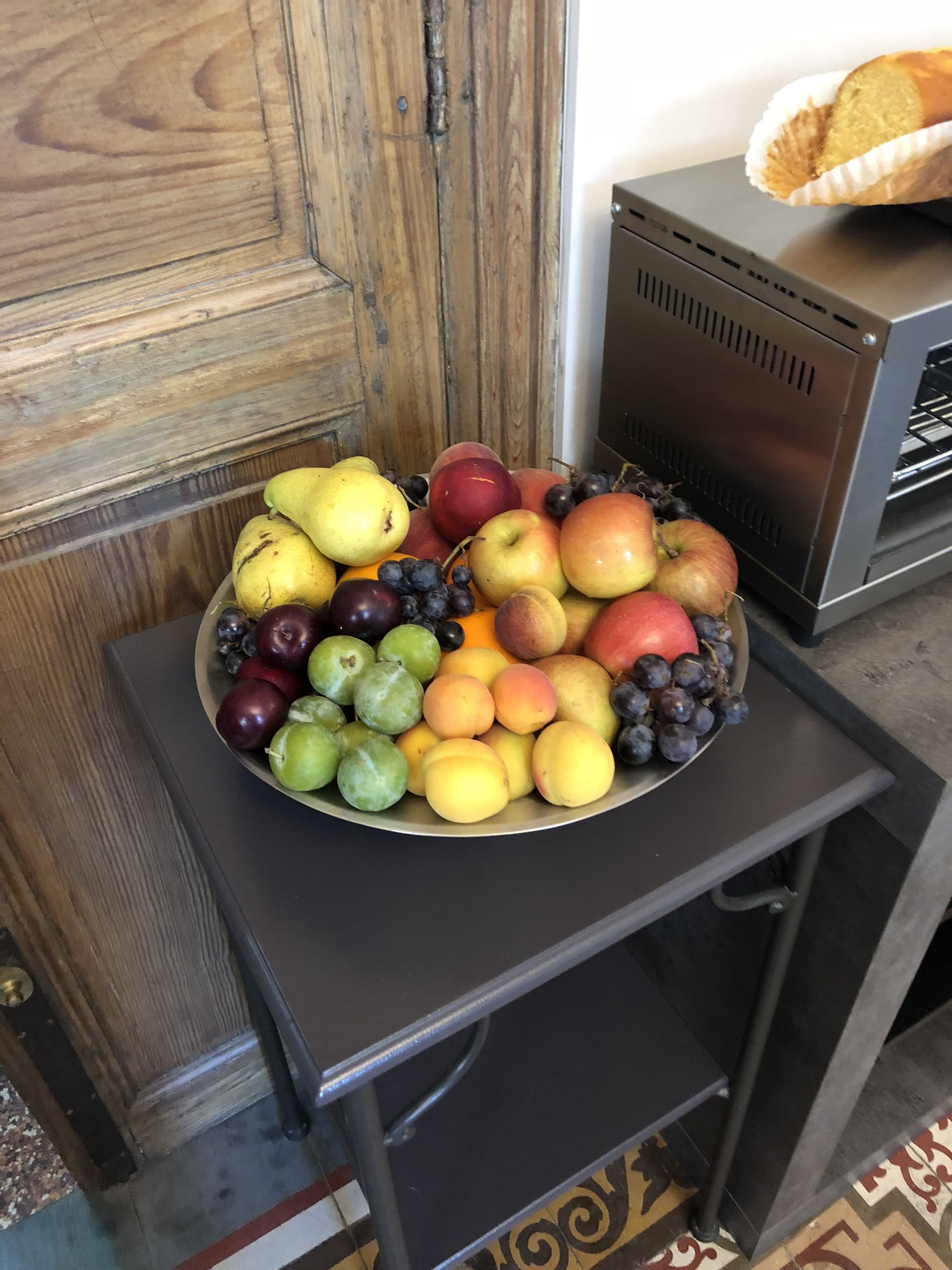 A fruit bowl on a table next to a wooden door and a bread box.