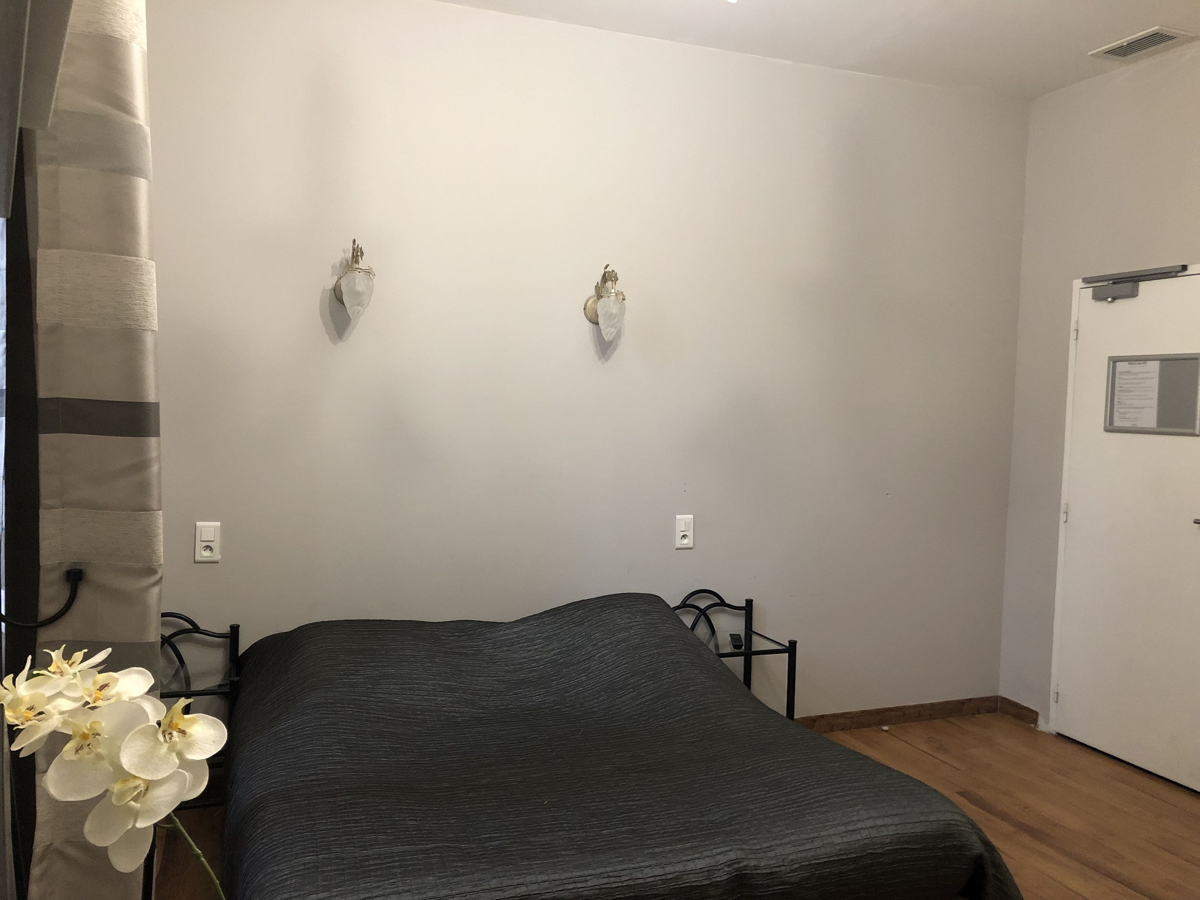 A bedroom with a bed, a nightstand, a wall-mounted light fixture, and a door with a notice.