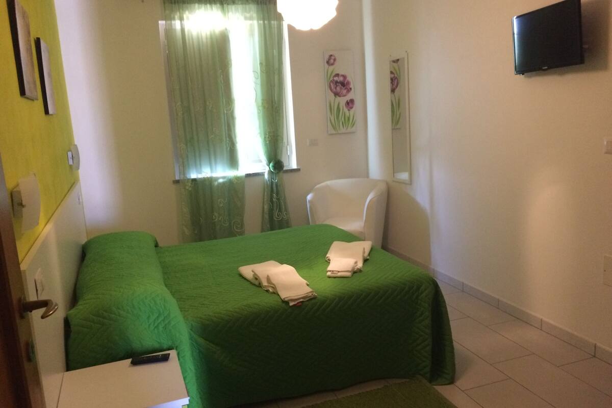 Una habitación con una cama verde, una silla blanca, un televisor de pantalla plana en la pared y una ventana con cortinas.