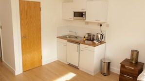 A compact kitchen with a wooden door, white cabinets, a microwave, a sink, and a small trash bin. - Trafalgar House (Stoke-on-Trent)