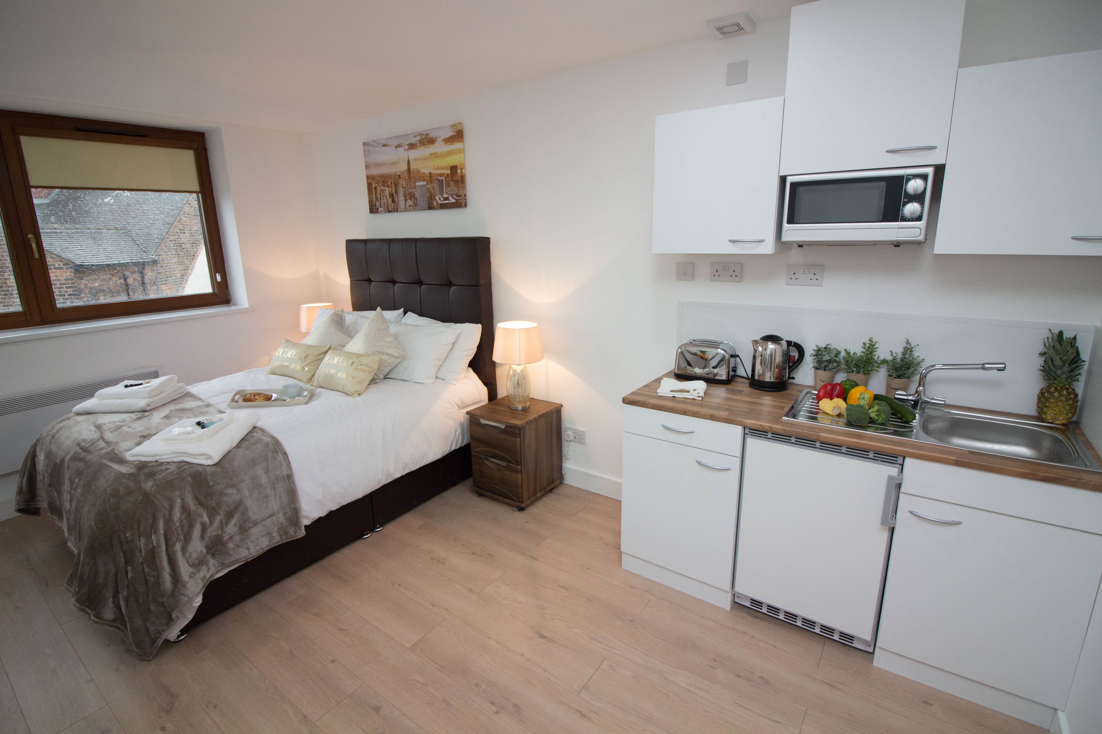 Ein Schlafzimmer mit Bett, Nachttisch und Ablagefläche. Im Hintergrund ist eine Küchenzeile mit Mikrowelle, Spüle und Schränken erkennbar.