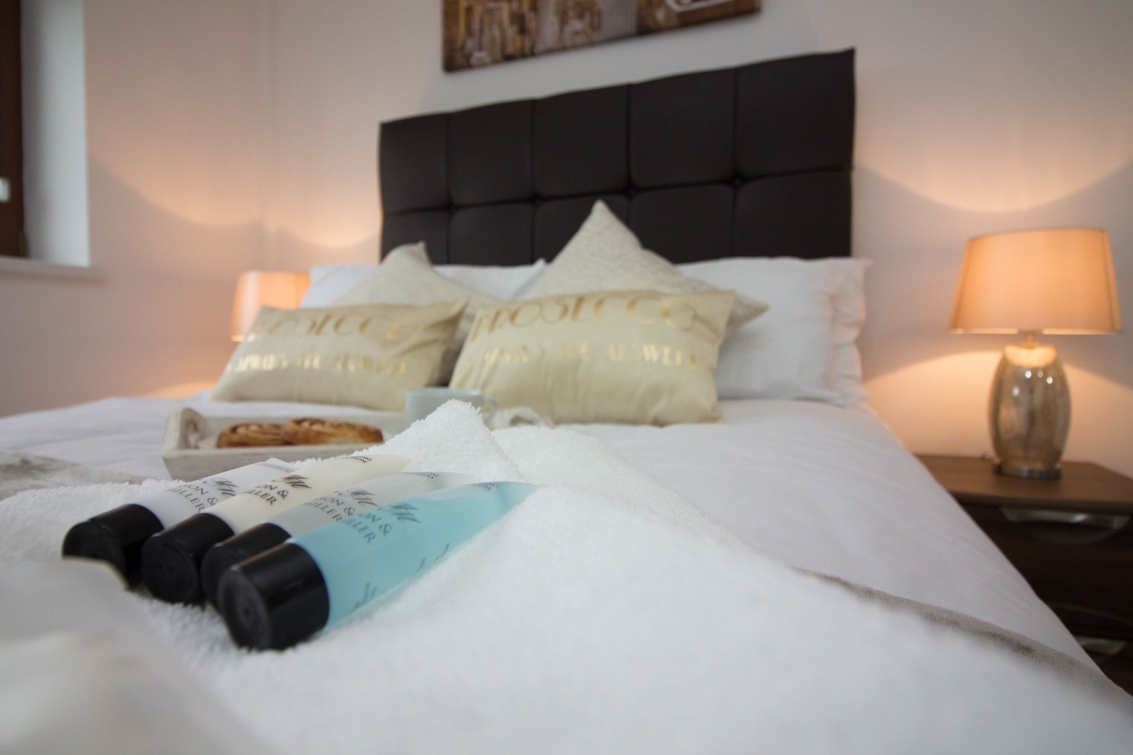 Un lit bien fait, avec du linge de lit blanc, une tête de lit noire et des coussins décoratifs ornés de motifs. On peut voir deux flacons de lotion sur le lit.