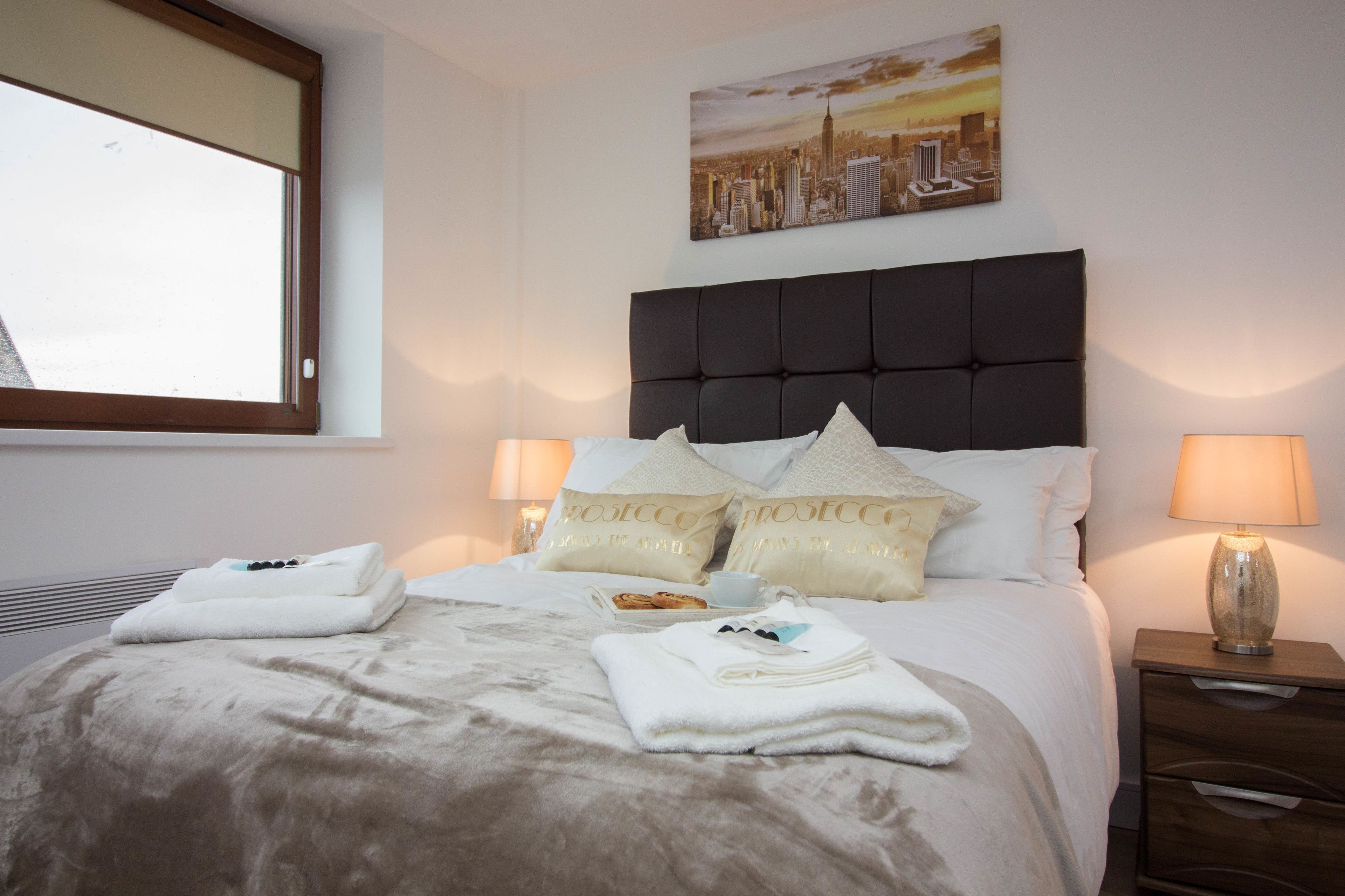 A neatly made bed with a cityscape painting above it, a bedside lamp, and a nightstand with a lamp.