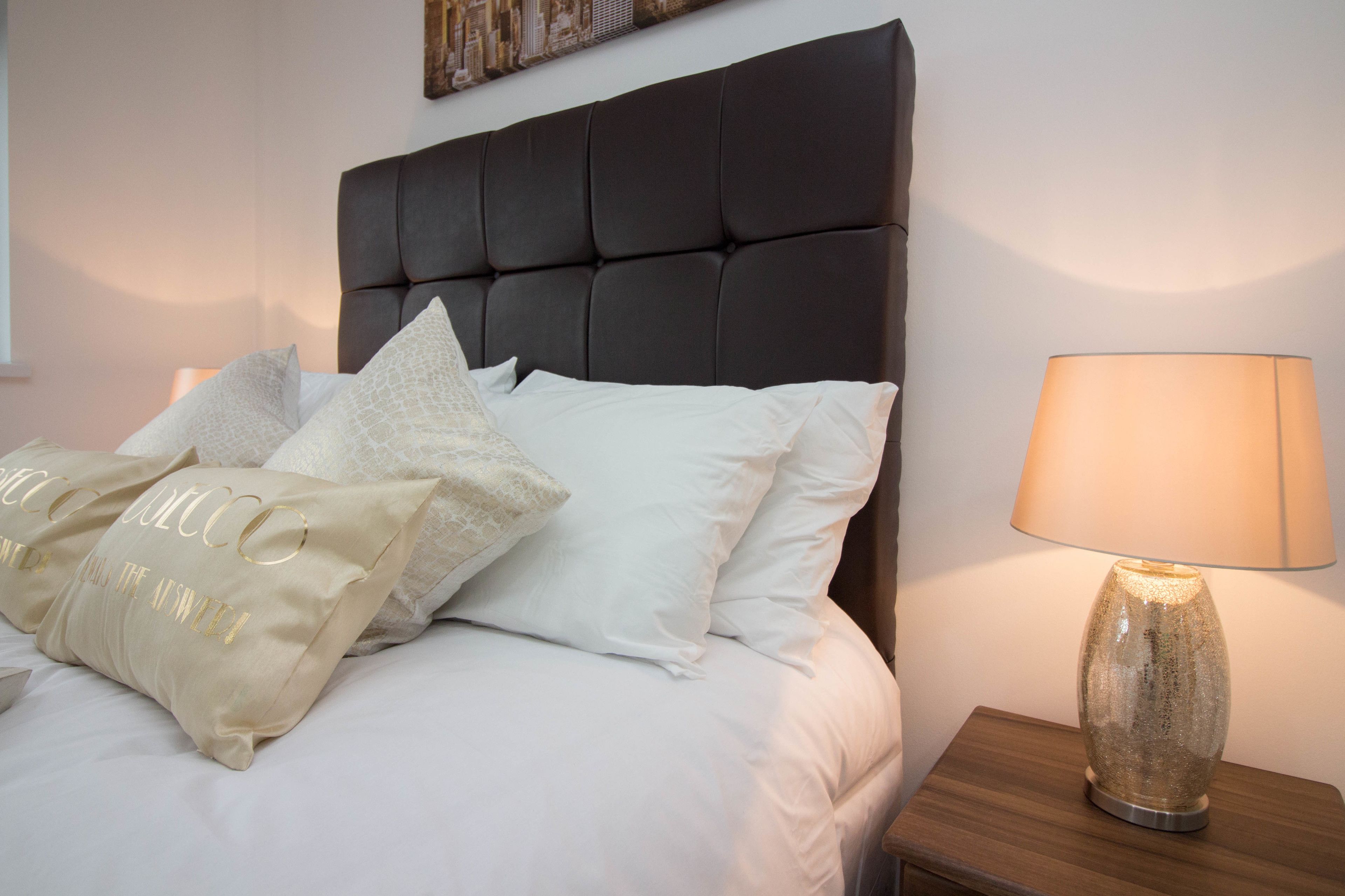Un letto rifatto con cura, lenzuola bianche e cuscini decorativi, una testiera imbottita scura e una lampada da comodino su un comodino in legno.