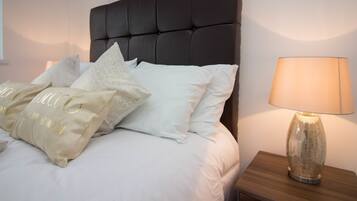 A neatly made bed with white linens and decorative pillows, a dark upholstered headboard, and a bedside lamp on a wooden nightstand.