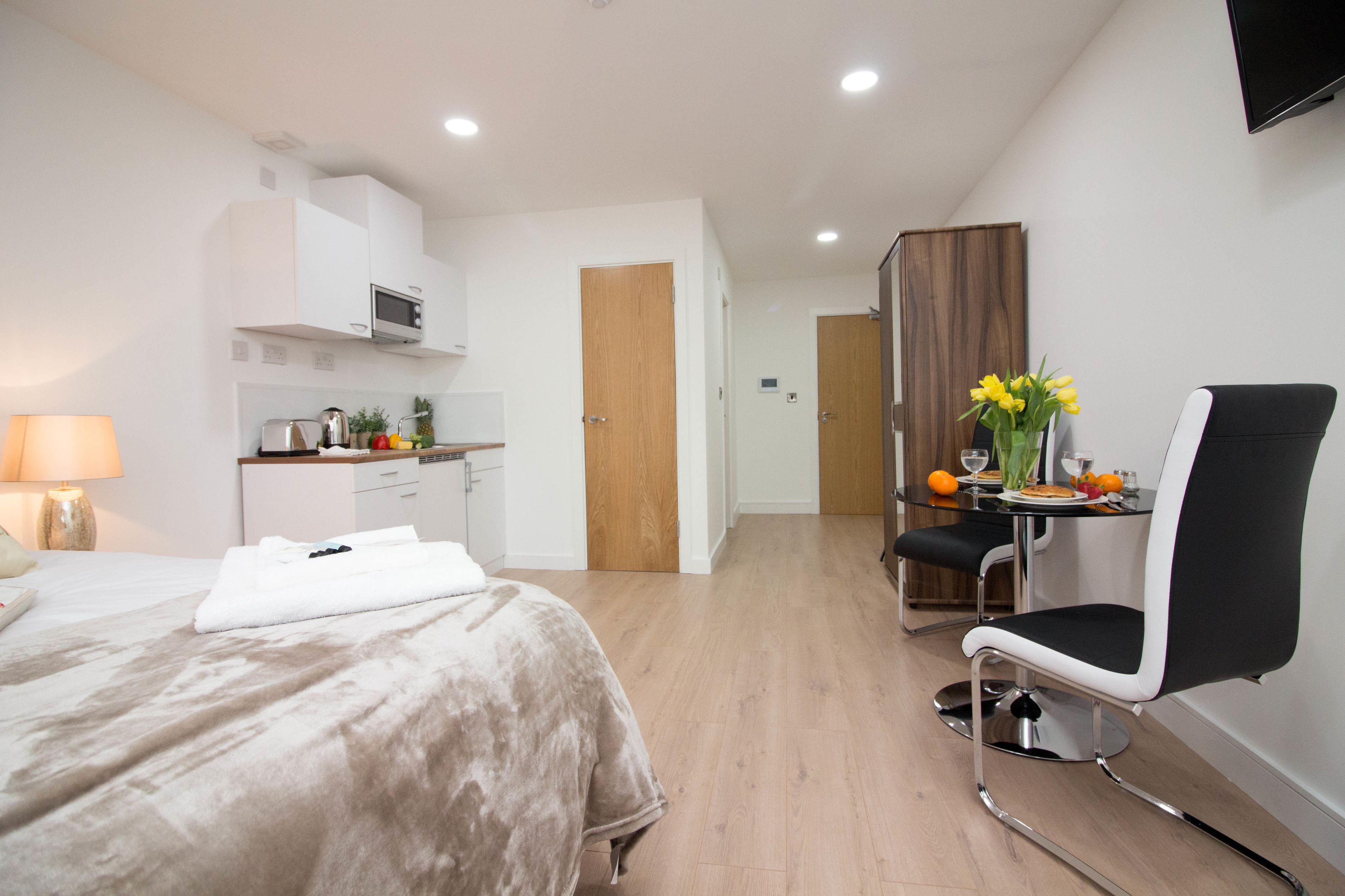 A modern hotel room with a bed, a small kitchenette, a dining area with a table and chair, and a TV mounted on the wall.