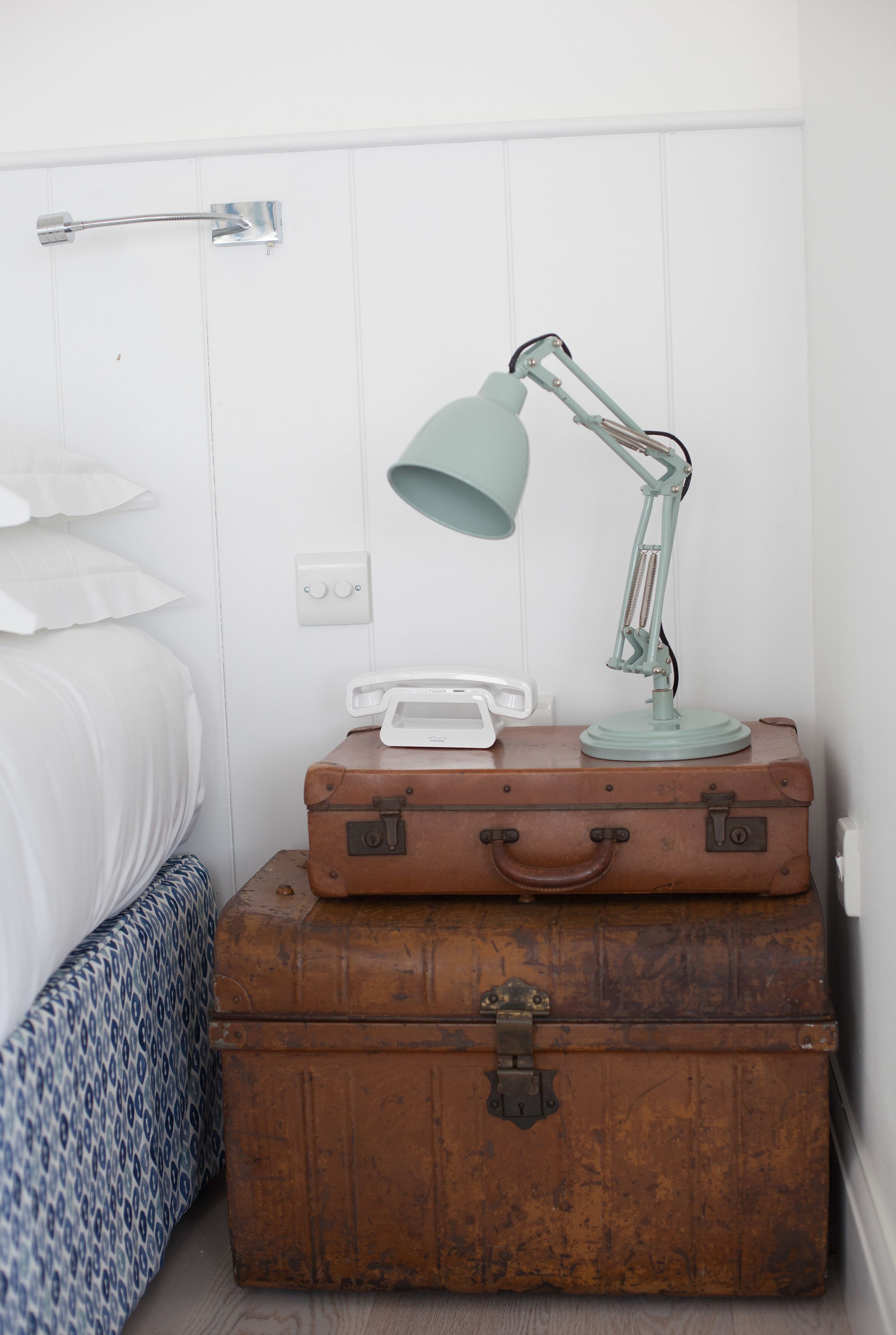 Een bed met witte lakens, een witte nachtkastje met een lamp, en een koffer erop.