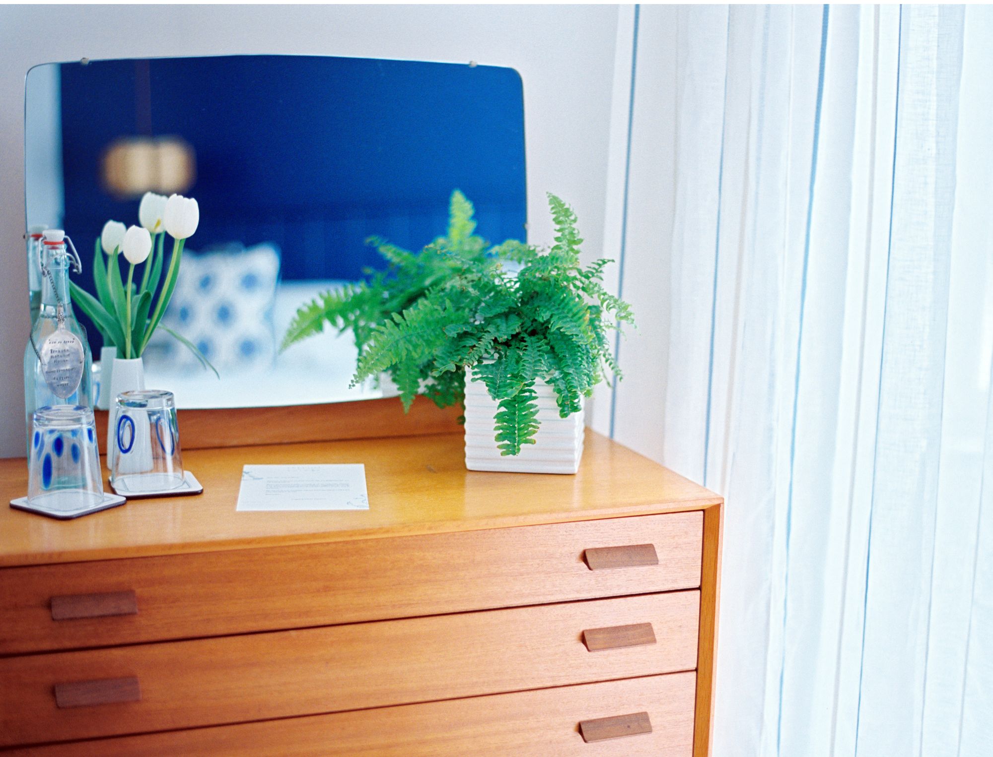 Een houten bureau met een kamerplant varens, een glazen fles en een vaas met witte bloemen.