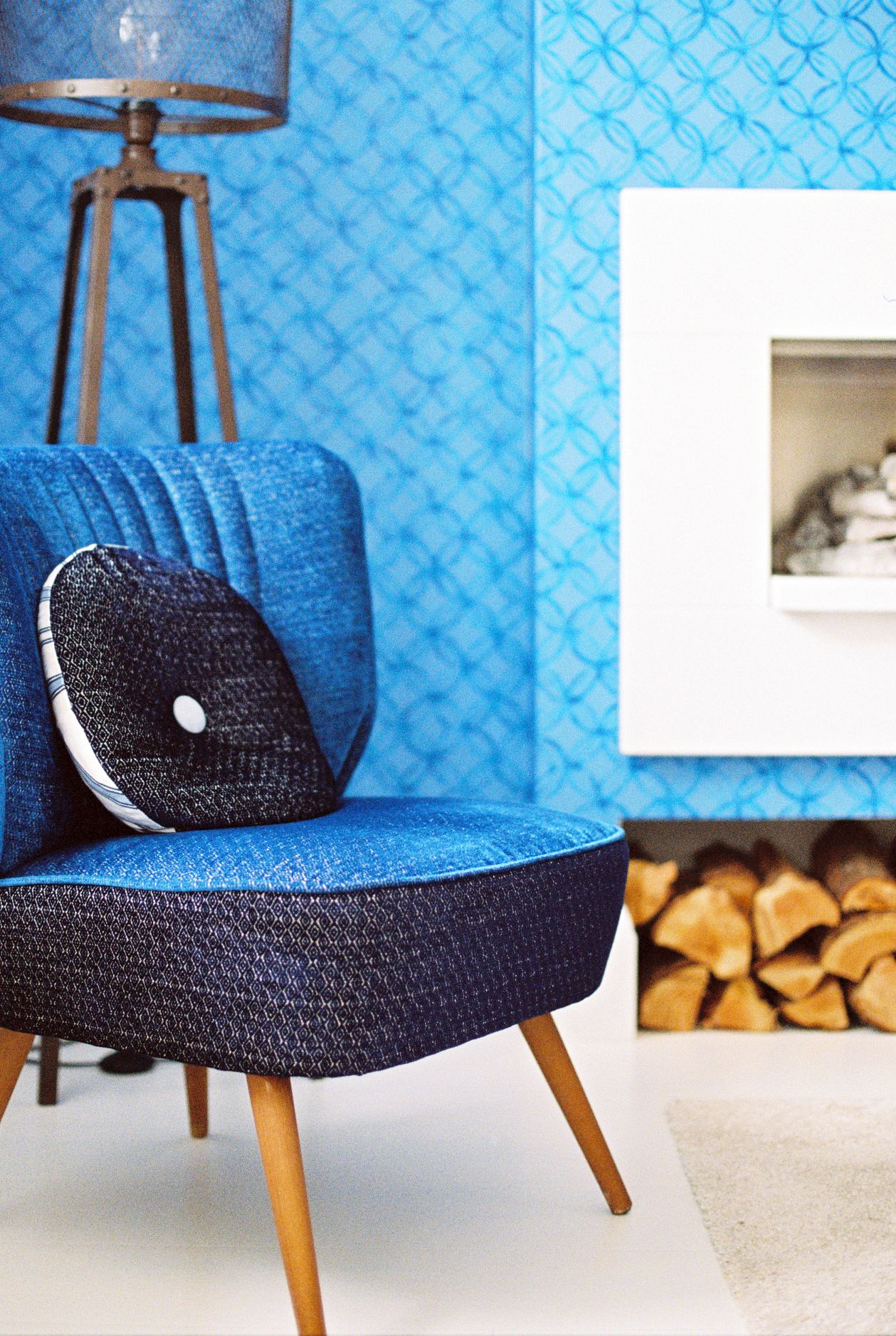 Un fauteuil bleu, revêtu d’un tissu texturé, avec un coussin noir, placé dans une pièce dont le papier peint est bleu à motifs et qui possède une cheminée blanche.