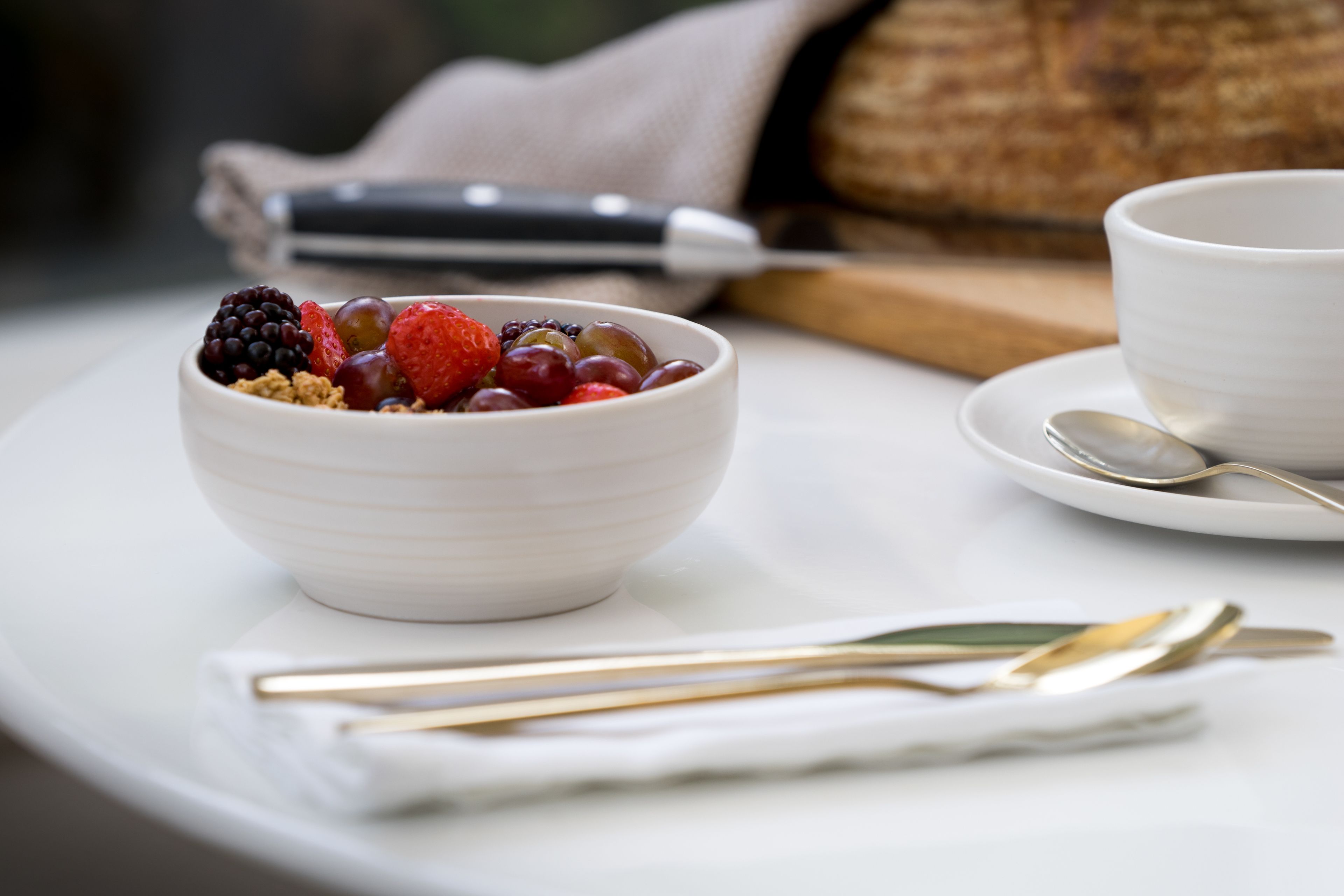 A white bowl with fruits and granola on a white plate, accompanied by a spoon and a knife on a napkin.