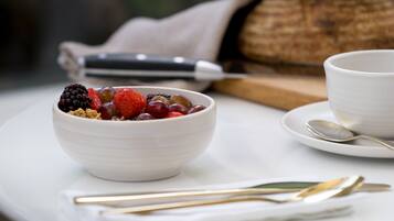 A white bowl with fruits and granola on a white plate, accompanied by a spoon and a knife on a napkin.