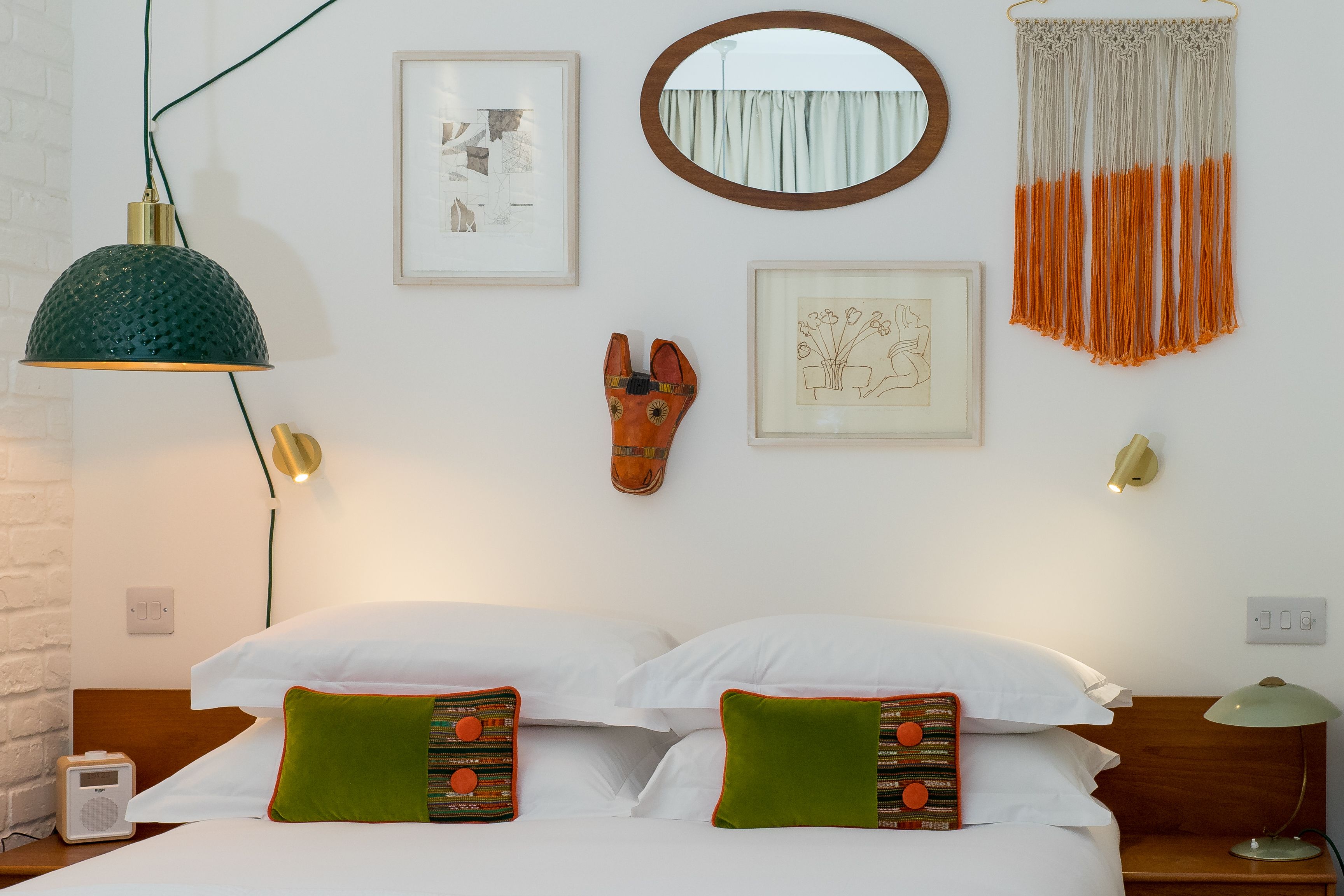 Una cama bien hecha con sábanas blancas y dos almohadas verdes, un espejo montado en la pared, una obra de arte enmarcada, un tapiz de macramé y una lámpara de noche.