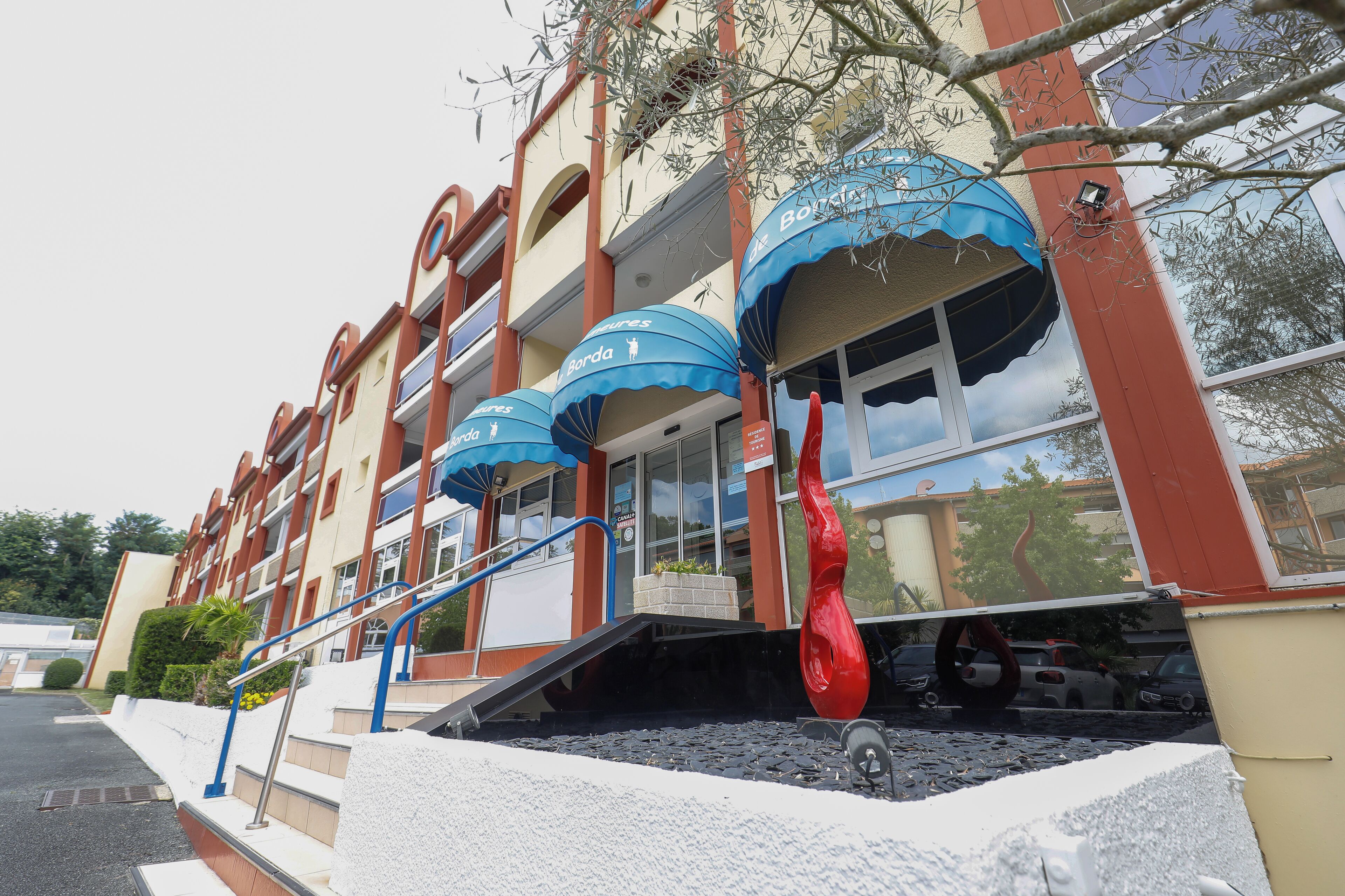 Un hotel con fachada roja y color beige, toldos azules y una escultura roja.