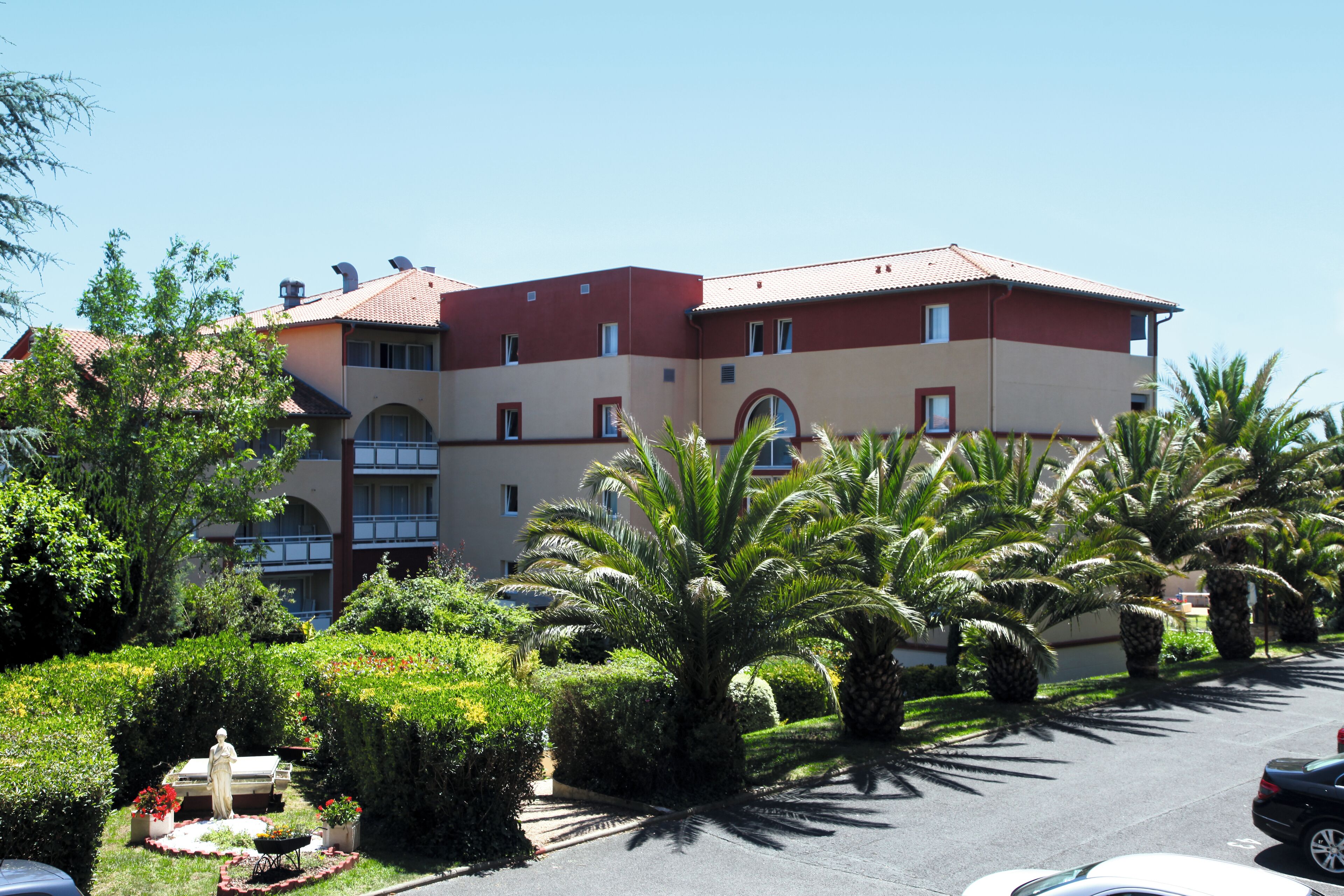 A multi-story hotel with a red and beige facade, surrounded by palm trees and well-maintained gardens.