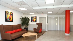 Un espace de bureau moderne avec un canapé marron, une table basse en bois et une chaise rouge. Deux grandes peintures ornent les murs, et une plante en pot est également présente.