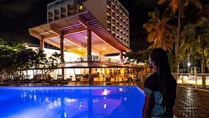 Zona de piscina con un área de descanso cubierta y un edificio al fondo.