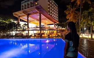 Zona de piscina con un área de descanso cubierta y un edificio al fondo.