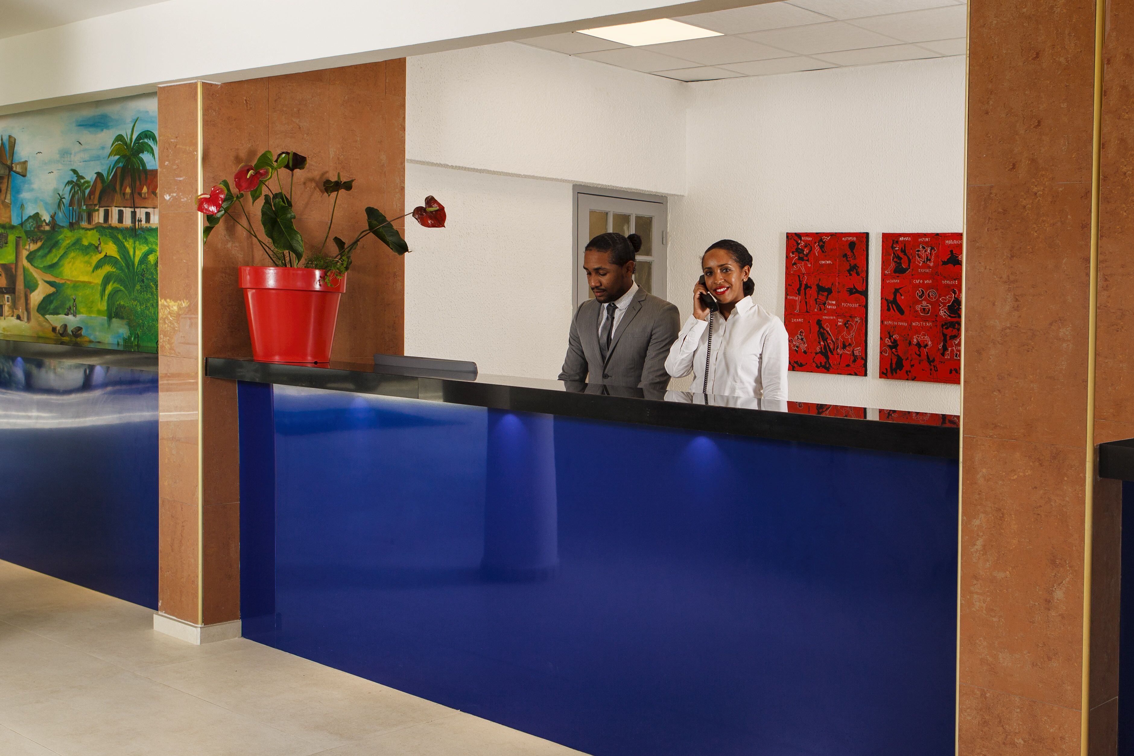 Un mostrador de recepción con una encimera azul, una maceta roja y un mural con un paisaje tropical.