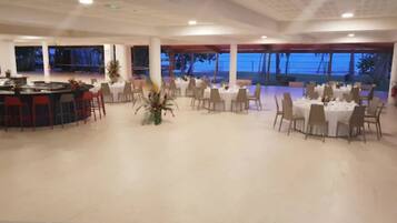 A spacious indoor area with multiple round tables and chairs, a bar area, and large windows offering a view of the outdoors.