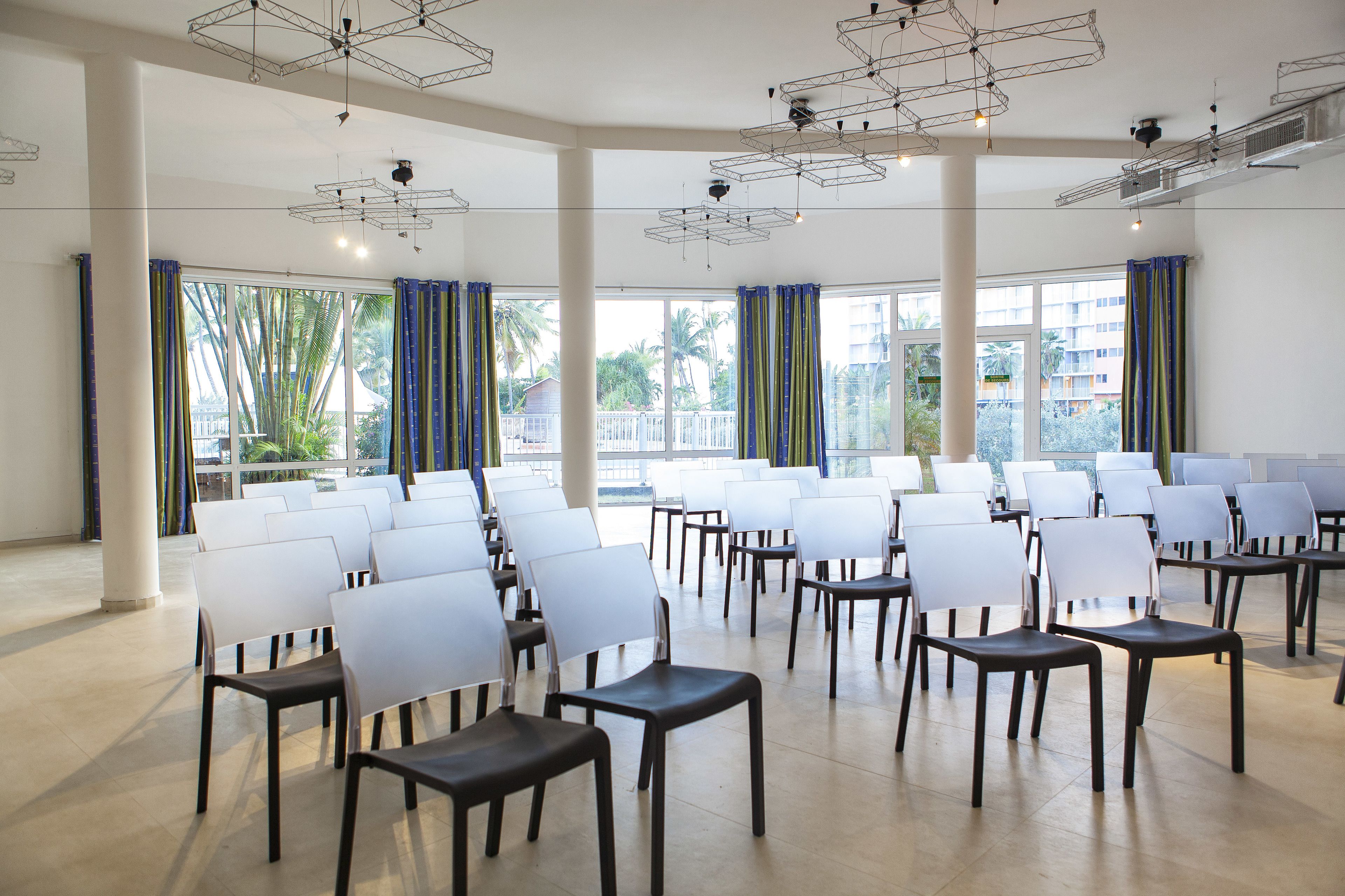 A conference room with rows of white chairs, large windows with curtains, and modern geometric ceiling lights.