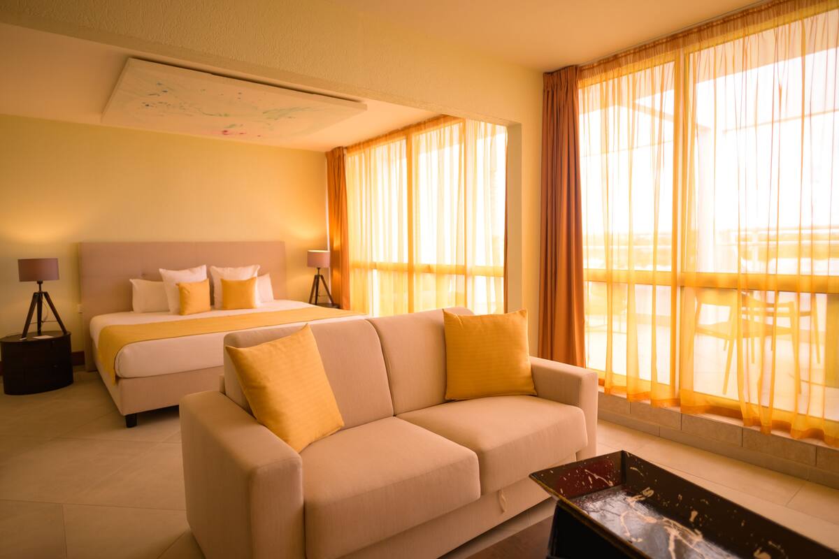 A hotel room with a bed, a sofa, a desk, and large windows with sheer curtains.
